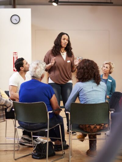 Woman Standing To Address Self Help Therapy Group Meeting In Community Center Woman Standing To Address Self Help Therapy Group Meeting In Community Center